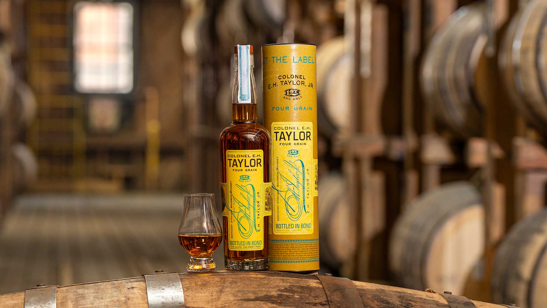 A bottle and glass of Colonel E.H. Taylor Four Grain bourbon sit on a barrel, with the bottle&rsquo;s yellow box beside them&mdash;an exceptional Father&rsquo;s Day gift. Wooden whiskey barrels line the blurred background inside a distillery warehouse.