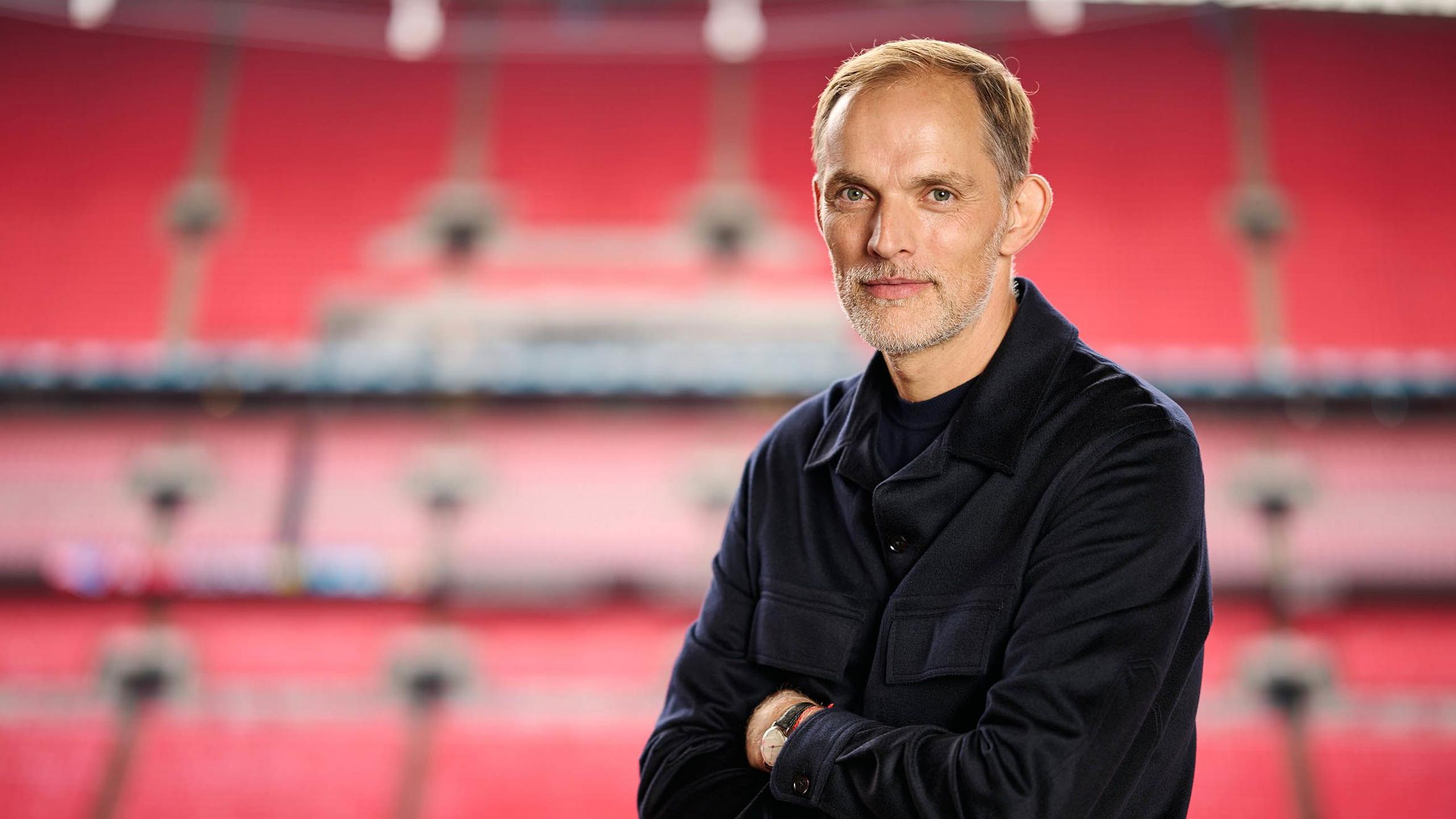 A man with short hair and a beard stands with arms crossed in a stadium.