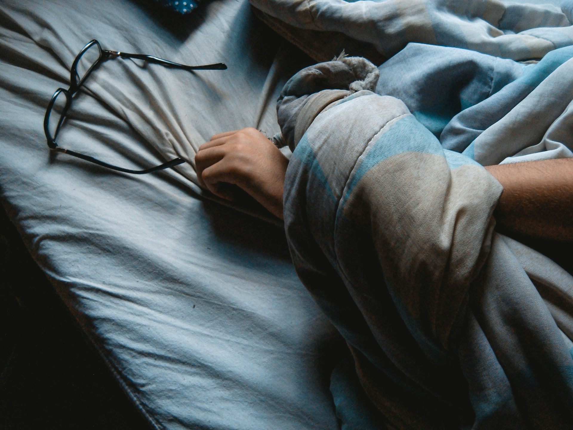 Arm entangled in bedsheets reaching towards a pair of glasses.